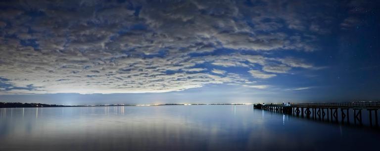 Mirjam van Sabben;Night Fishing at Cornwallis;This photo was taken on the 9th of June 2018 at Cornwallis beach. I hadn’t intended to take photos at this location but on a whim decided to swing by it on my way to Little Huia. My impromptu decision rewarded me with a spectacular view of a large cloud formation over the Manukau Harbour. Stars peek through its gaps. On the left under the glow of the city, the top of the Sky Tower and its reflection on the water can be seen. To the right, people are fishing from the wharf. I wasn’t confident I would be get them looking sharp with my long exposure, but one fisherman held still for the entirety of my shot, beautifully silhouetted against the night sky.