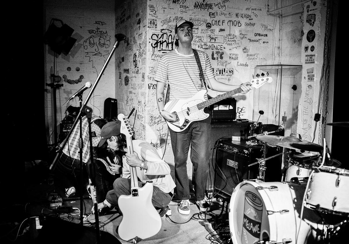 David Dunham; FESHH, Underground, Auckland;Aesthetically, The Underground was rad, largely in part to it being an old, gigantic vault. The door appeared original in that you would need dynamite to prise it open, and there was no natural light. To the left was a burgundy leather sofa from a cigar era, the stage was not a stage but just a power cord lined section at the back of the room, and the ceiling was flanked by the structural foundations of the arcade. It was solid, felt air raid safe, and was square box shaped which meant noise bounced around with no nook and cranny to escape except for the bathroom that was next to the ‘stage’ and was reached by sometimes having to step onto the stage. Spending a penny came at the cost of feeling awkward.  When a band had more than three people, things got cramped, hence why in the picture above you’ll see the guitarist for FESHH playing statue as his bandmates prepare for their set as part of The Others Way Festival.
